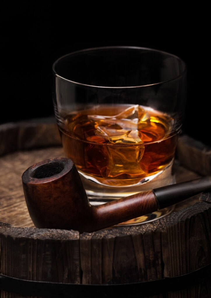 Glass of whiskey with ice cubes and vintage smoking pipe on top of wooden barrel.